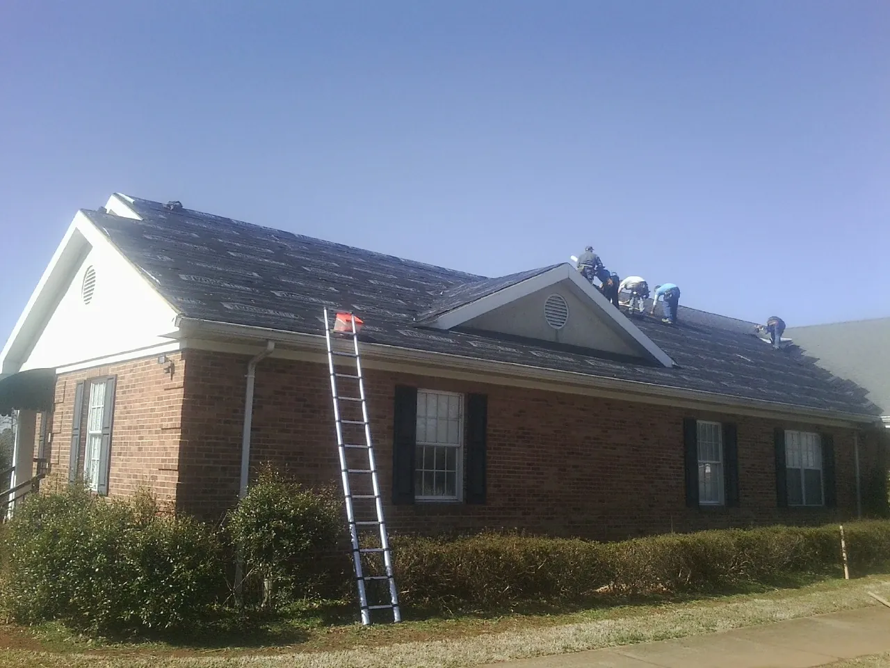 Crew tearing shingles off a brick ranch roof with a ladder set — Emergency Roof Repair work by our crew in Normal