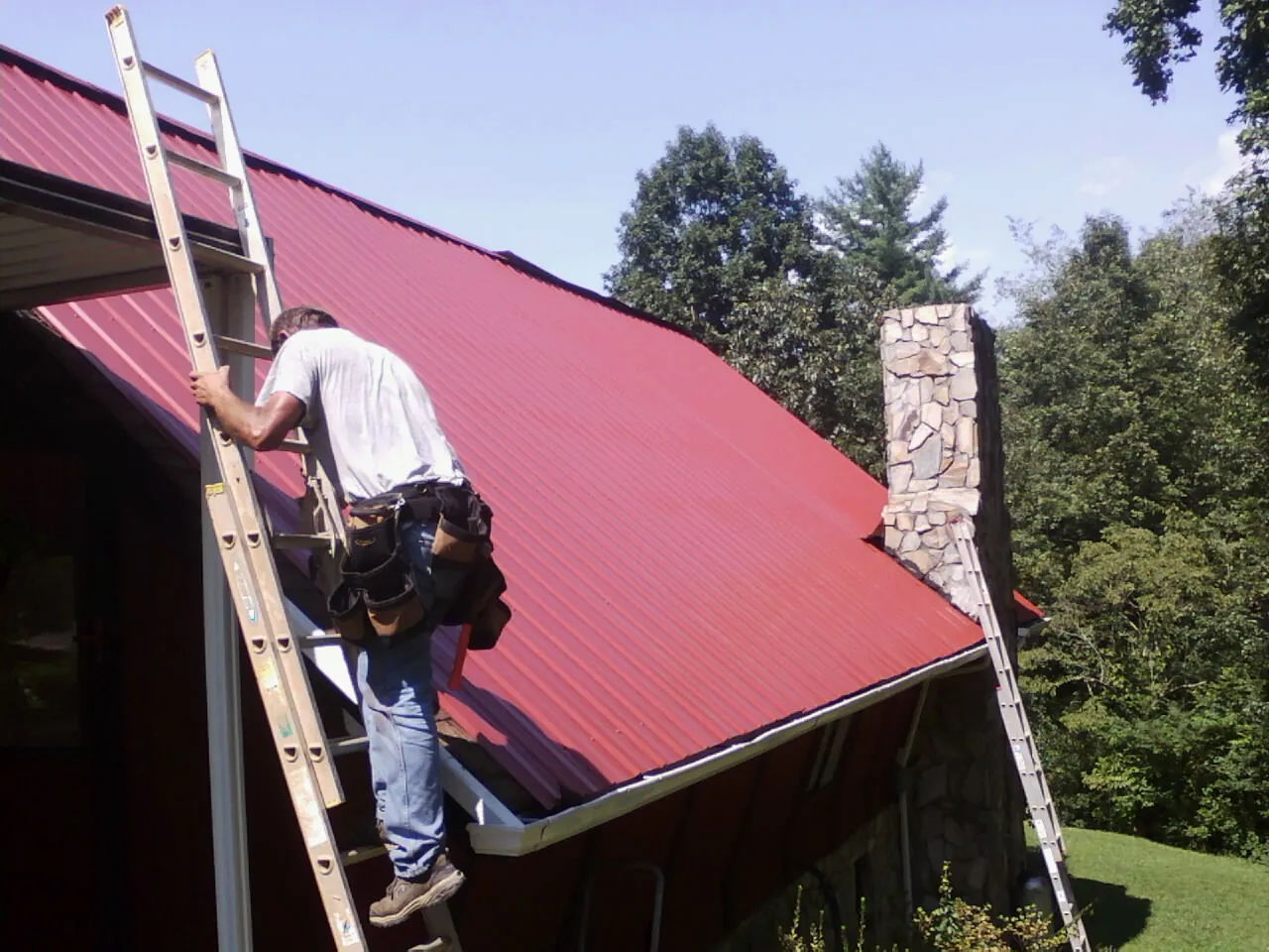 Roofer on a ladder beside a red metal roof and stone chimney — Soffit & Fascia Repair by Roofing Brothers in Normal